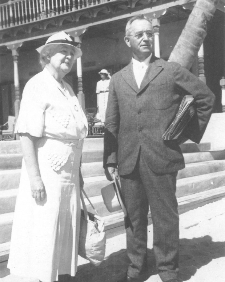 Evelyn Grove Seely, in a white dress and hat, stands beside her husband Fred Seely, who wears a suit and holds a wide-brimmed hat in his hand.
